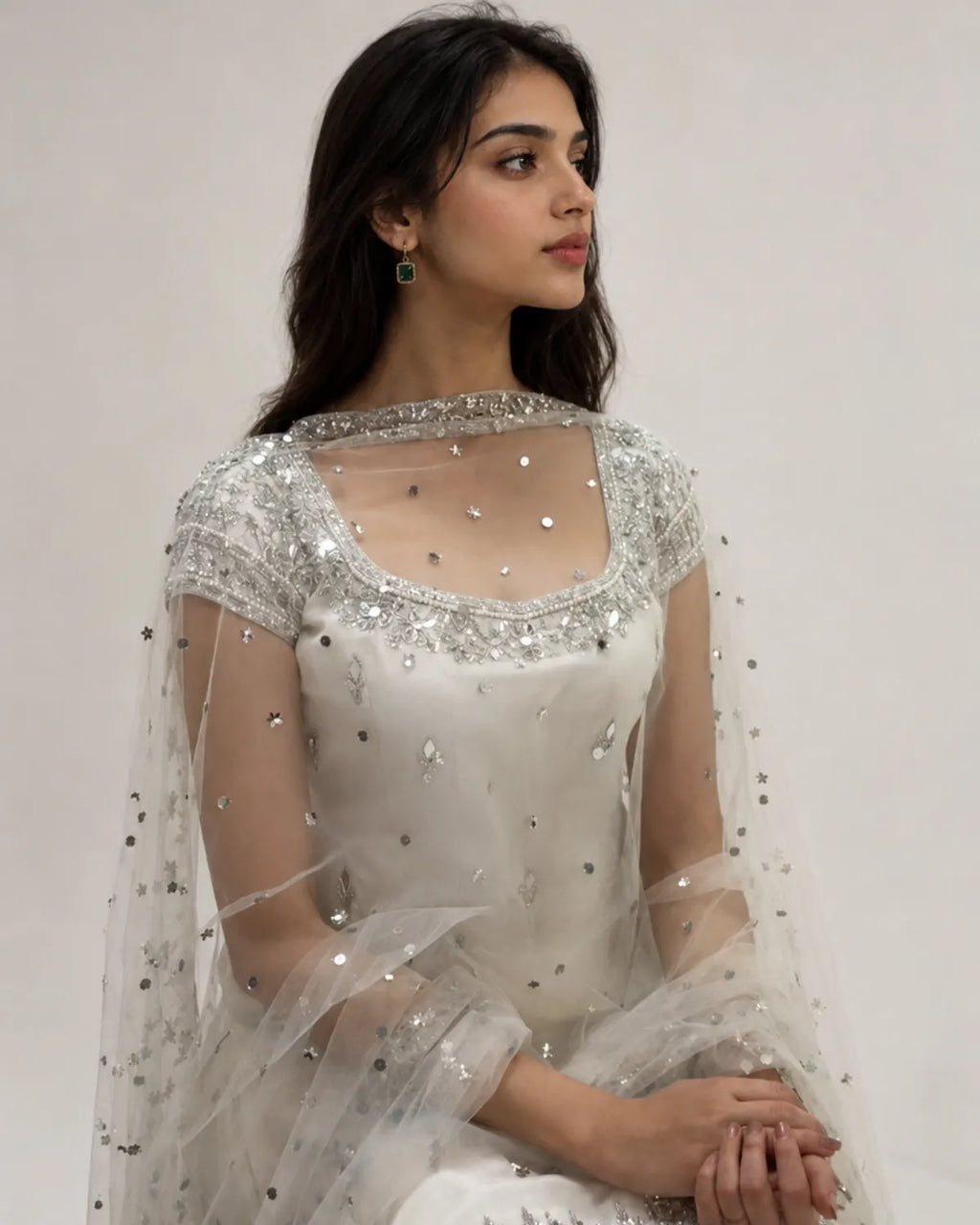 A woman wearing a white embellished traditional outfit with a sheer embroidered cape, photographed against a plain light background.