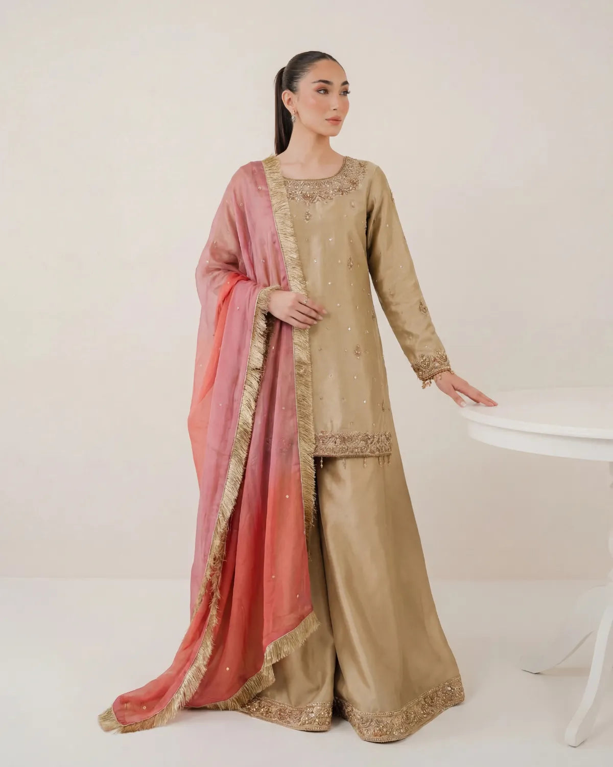 A woman models a traditional South Asian outfit consisting of a beige kurta and wide-leg trousers with gold embroidery, paired with a pink dupatta featuring a gold border.