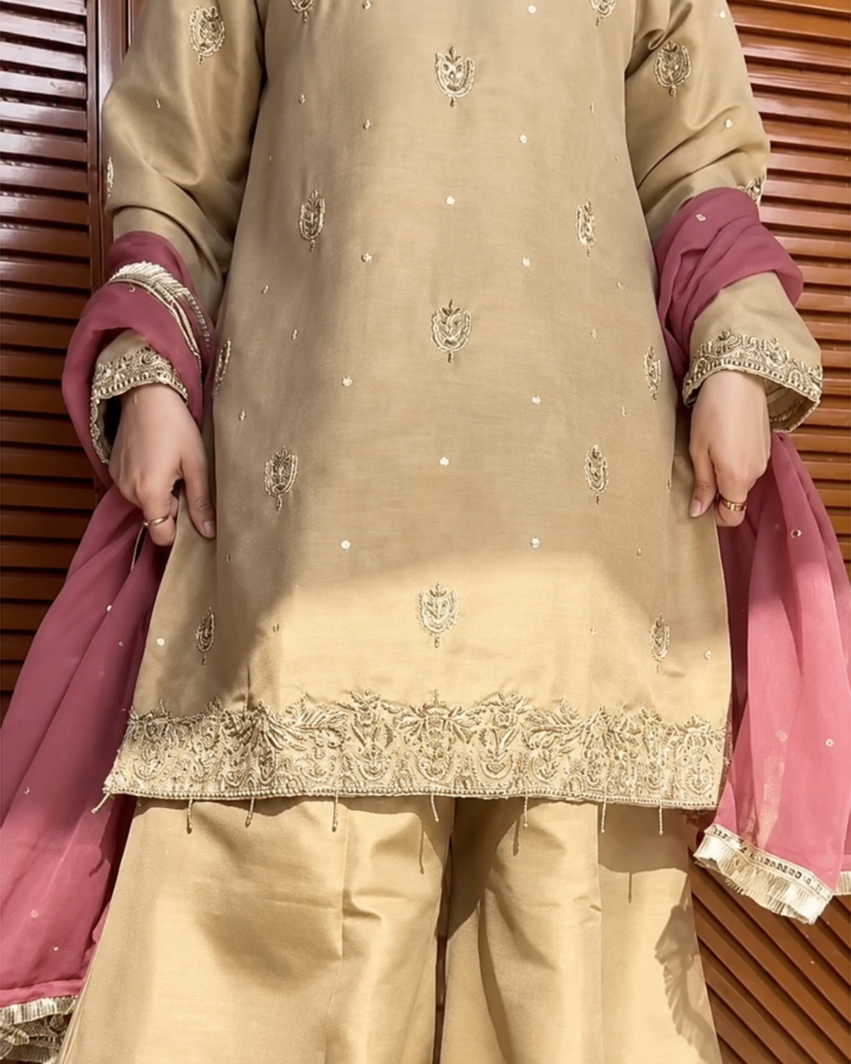 A close-up of a beige embroidered kurta with matching beige trousers and a pink dupatta draped over the arms, set against a brown wooden slat background.
