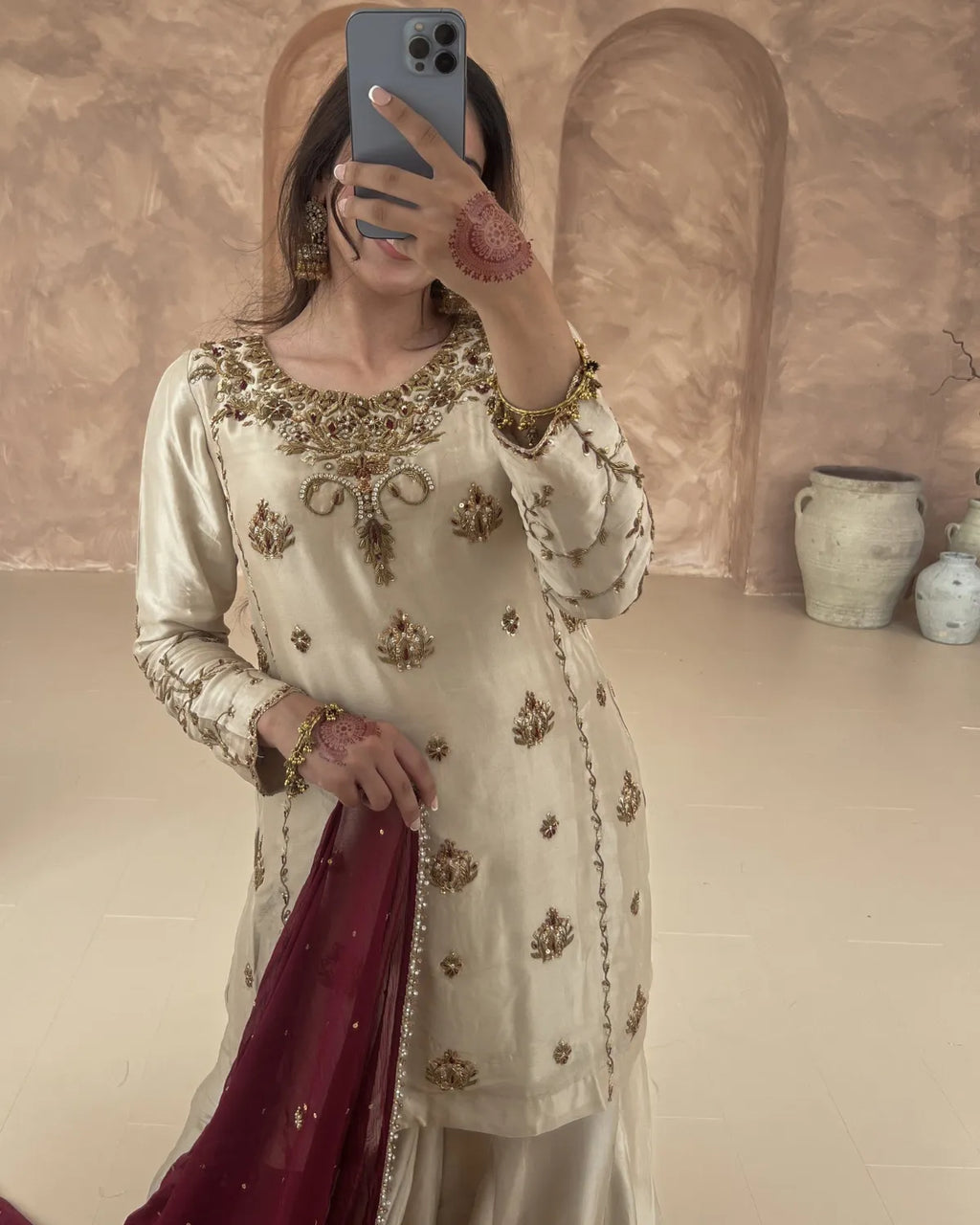 Person wearing a cream embroidered outfit with gold details, holding a phone and a maroon fabric.