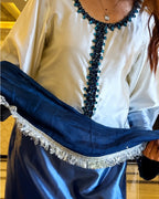 A close-up of a person wearing a white satin-like tunic with a dark blue beaded neckline and holding a matching dark blue draped fabric with silver fringe.
