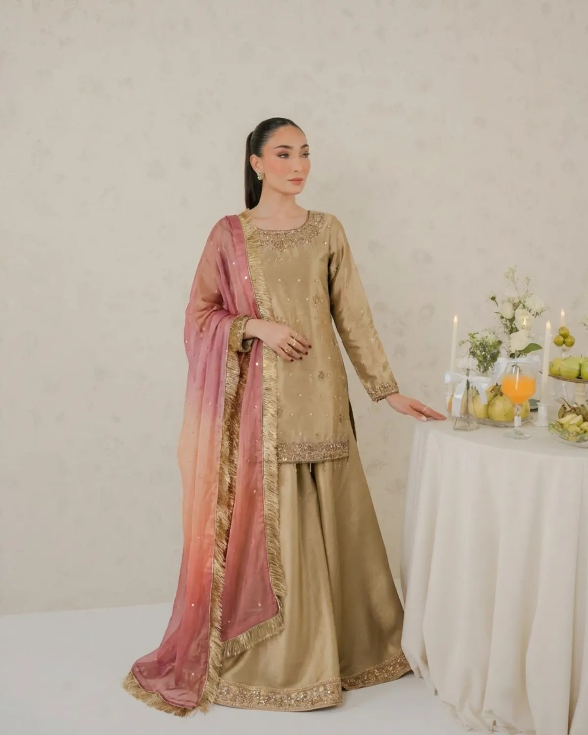 Woman in traditional beige and pink outfit standing next to a table with fruits and candles.