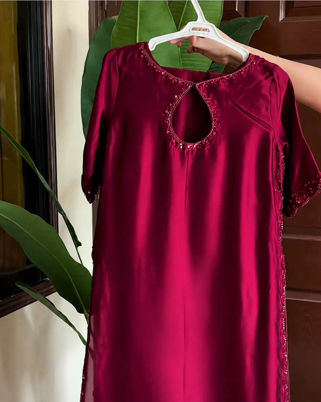 A deep magenta-red satin nightgown with a keyhole cutout and embroidered details, held up on a white hanger by a hand in front of a background of green leaves and a wooden door.