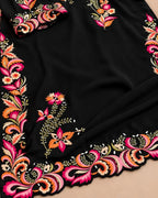 A close-up of a black fabric garment or textile featuring ornate multicolor floral and paisley embroidery along the border, with a matching embroidered cuff or sleeve detail visible at the top left.