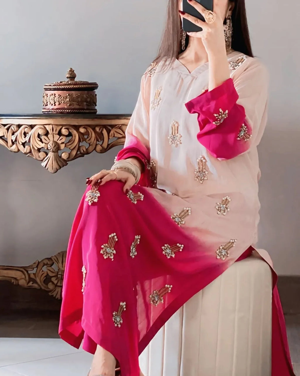 Person wearing a pink and beige saree with gold embroidery, sitting on a chair.