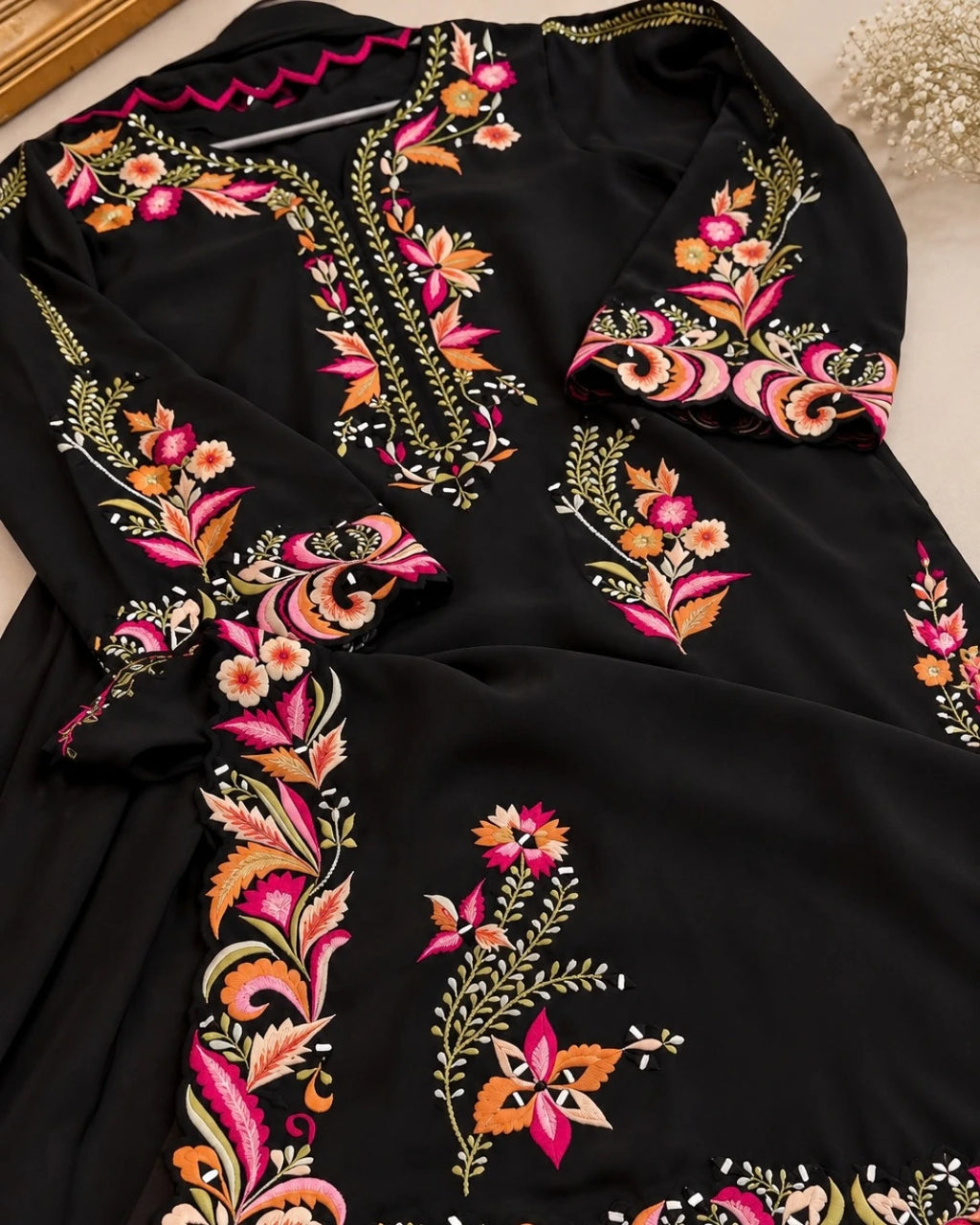 A black long kaftan dress featuring vibrant floral and paisley embroidery in pink, orange, gold, and green, laid flat on a light beige surface.
