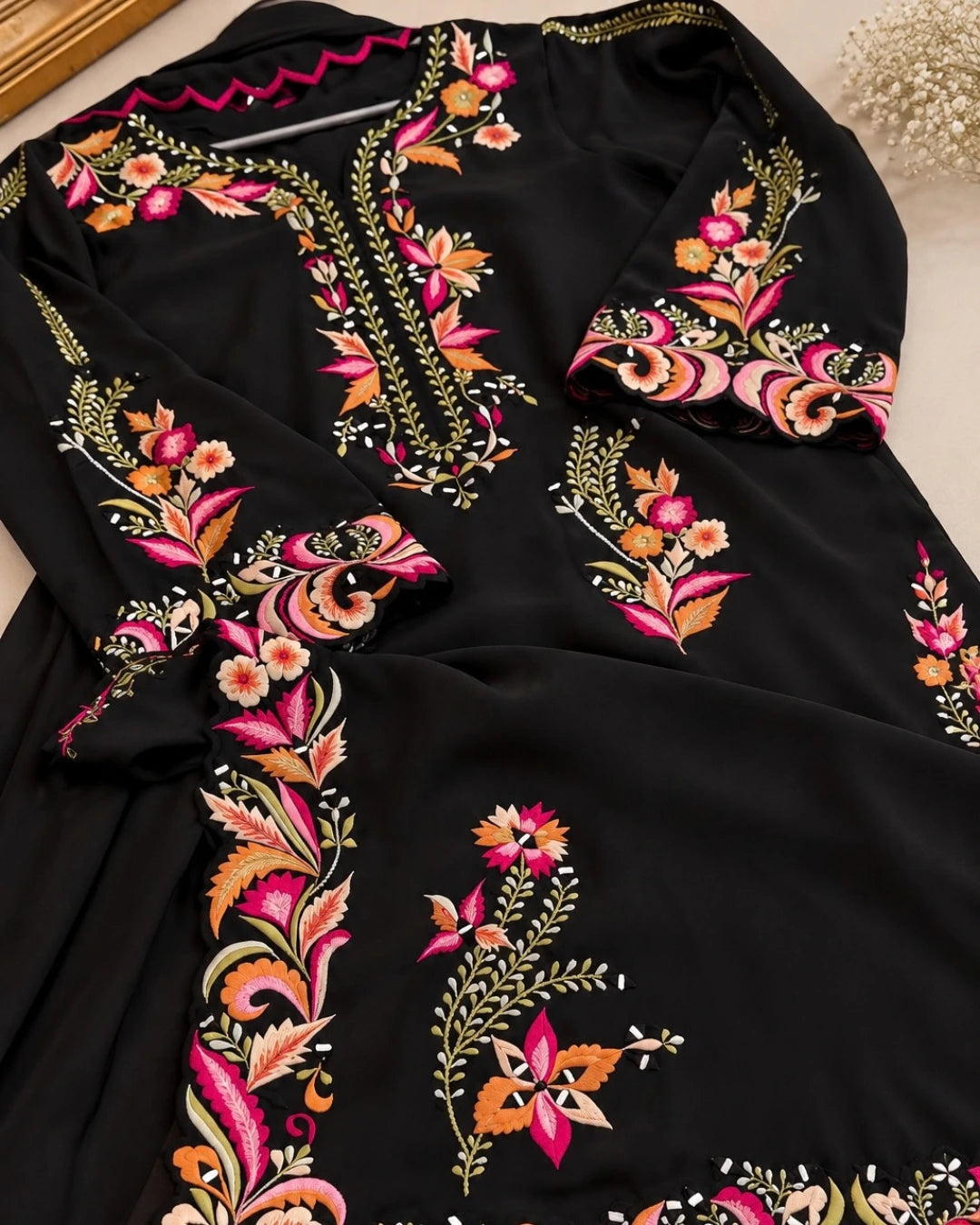 A black long kaftan dress featuring vibrant floral and paisley embroidery in pink, orange, gold, and green, laid flat on a light beige surface.