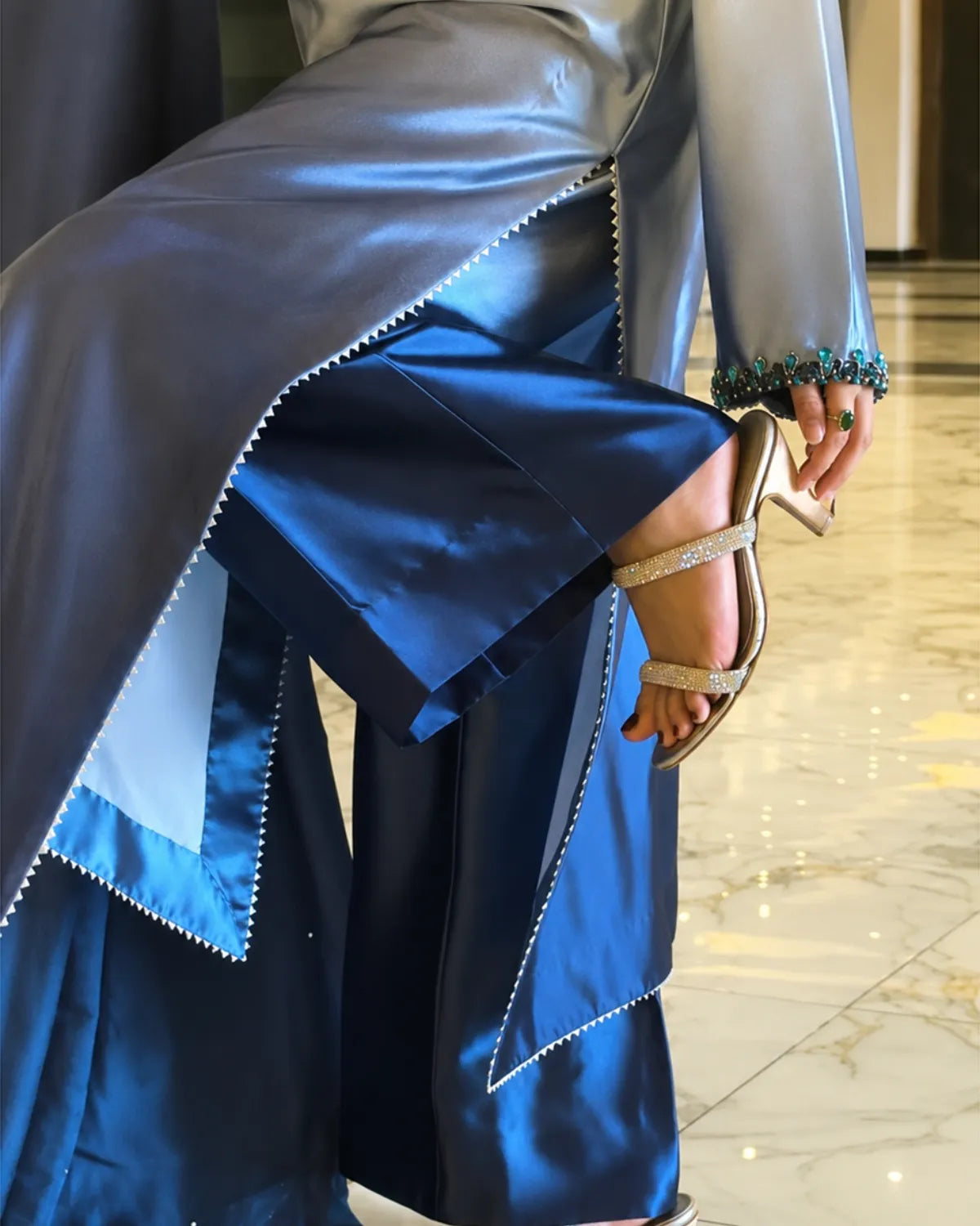 A close-up of a person wearing a flowing blue satin garment with layered flared panels and silver embellished sandals, standing in a polished indoor space with marble flooring.