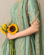 QAM Style luxury ethnic wear showcasing a sage green silk outfit with intricate golden embroidery, ornate borders, and a graceful traditional silhouette.
