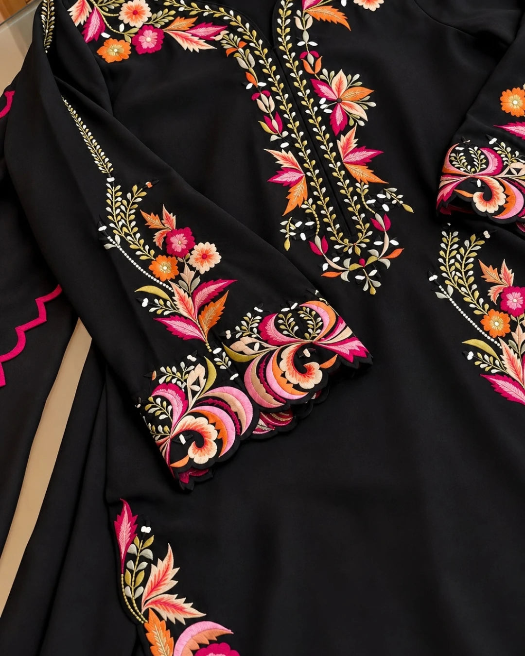 Close-up of a black embroidered garment, likely a kurta or dress, featuring intricate floral and vine embroidery in pink, orange, gold, and white on the neckline and sleeve cuffs.