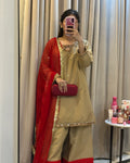 A woman takes a mirror selfie wearing a beige and red traditional Indian outfit with gold embroidery, holding a red clutch bag. The background shows beige curtains and a small table with cosmetics.
