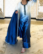 A woman wearing a blue satin traditional outfit with an ombré effect, featuring a white-to-blue gradient tunic with dark blue embroidery and matching wide-leg pants, holding a coordinating blue dupatta in an indoor setting with marble floors.
