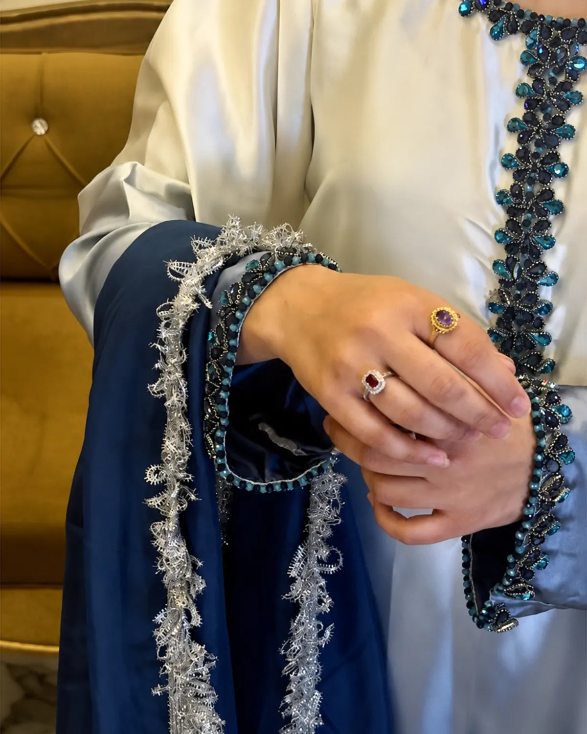 A close-up of a person wearing a cream satin robe with dark blue and teal beaded trim and a navy blue draped fabric with silver fringe, while wearing two rings on their fingers.