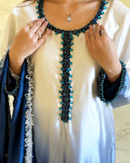 A close-up of a woman wearing a white satin tunic with elaborate dark blue and teal beaded embroidery around the neckline and cuffs, paired with a matching blue draped shawl.