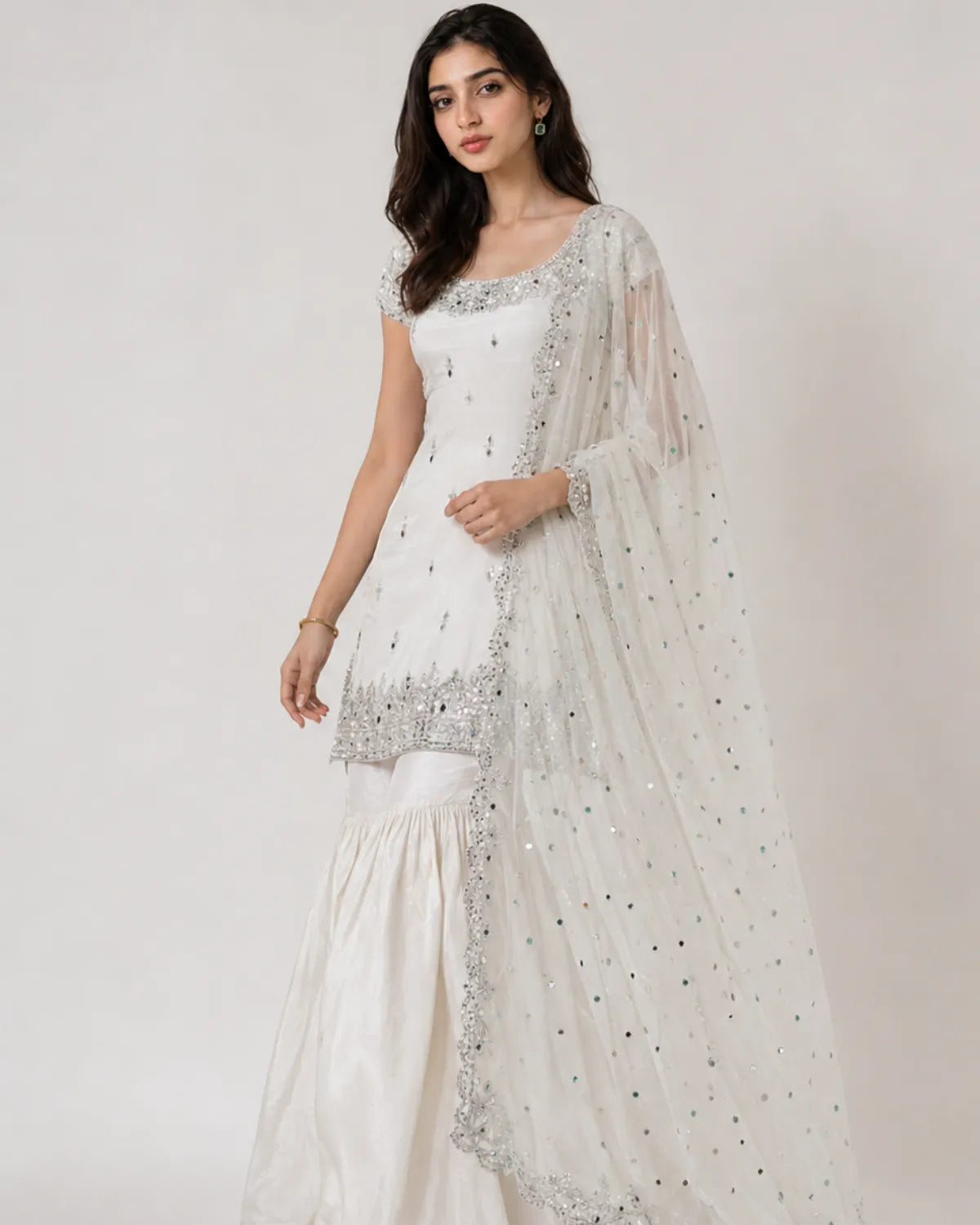 A woman modeling a white traditional outfit with silver embellishments and a sheer white dupatta with scattered silver mirror work, photographed against a plain light background.