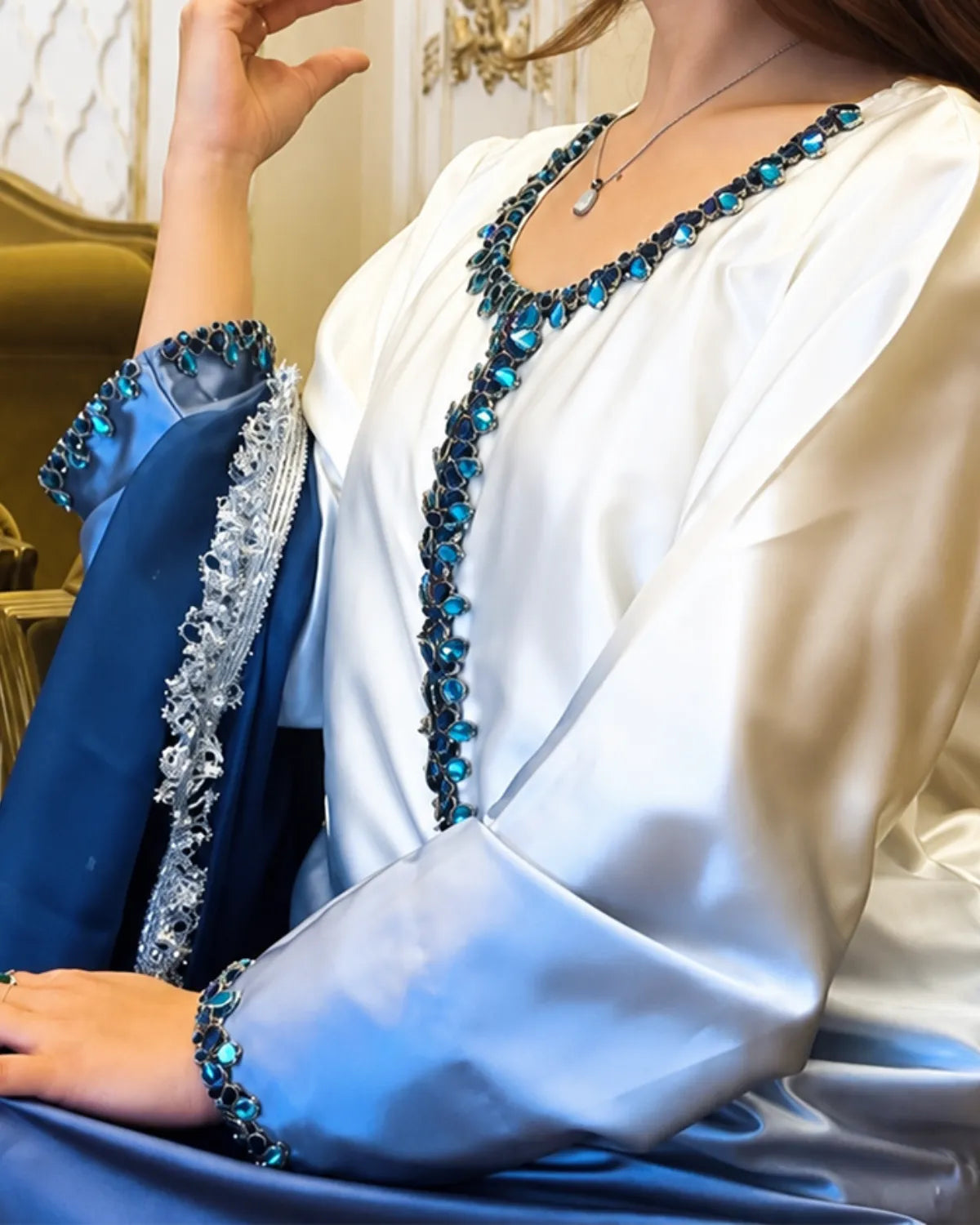 A close-up of a woman wearing a white satin robe with a blue embroidered border and matching blue draped fabric, featuring sparkling blue rhinestone embellishments along the neckline and sleeve cuffs.