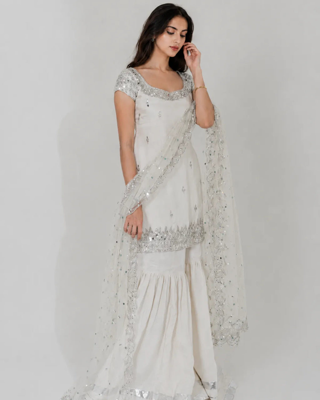 A woman modeling a traditional white embroidered salwar kameez set with a matching sheer dupatta, featuring silver sequin and bead embellishments, against a plain light gray background.