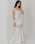 A woman modeling a traditional white embroidered salwar kameez set with a matching sheer dupatta, featuring silver sequin and bead embellishments, against a plain light gray background.