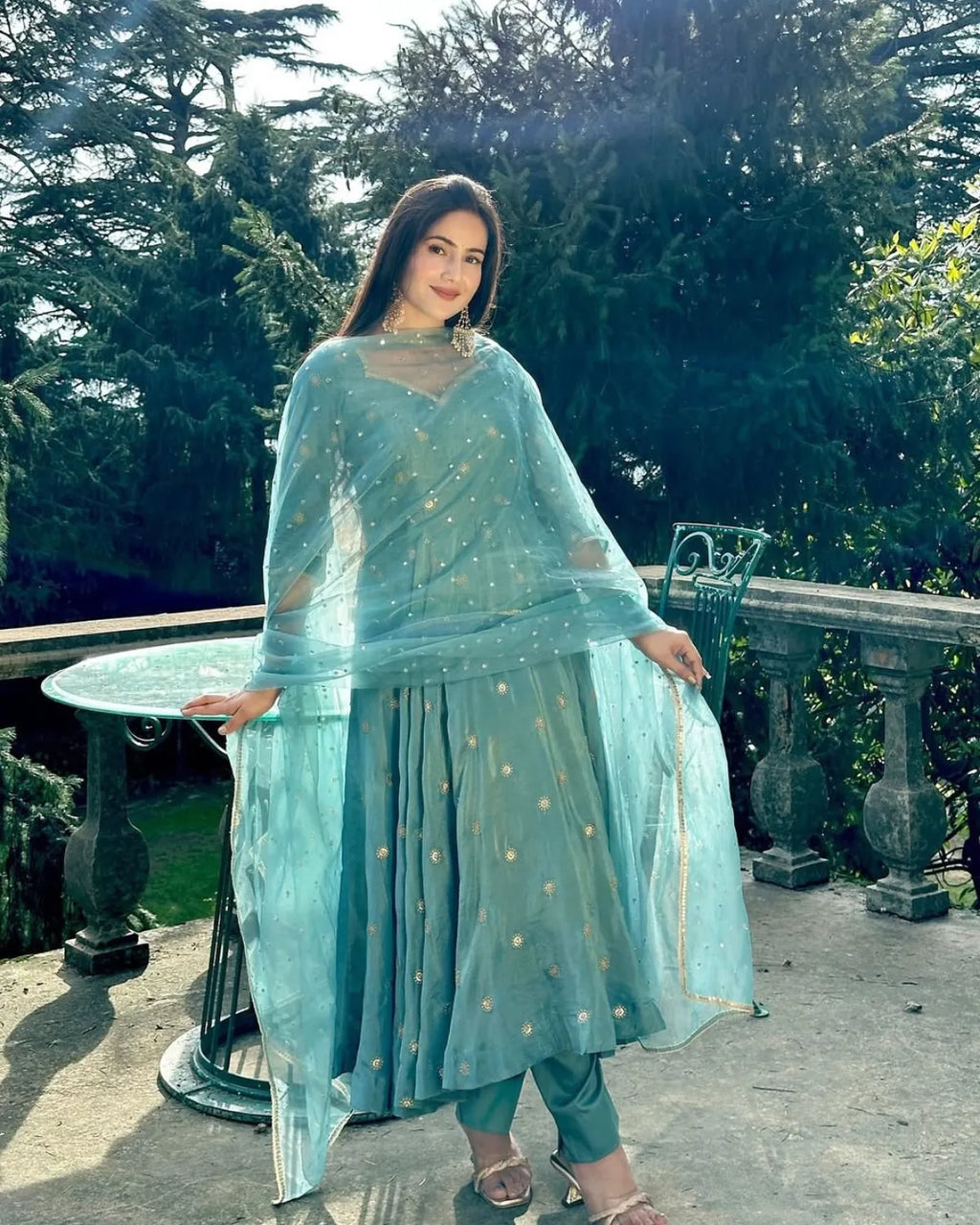 Woman in a light blue traditional outfit standing outdoors with greenery in the background