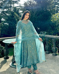 Woman in a light blue traditional outfit standing outdoors with greenery in the background