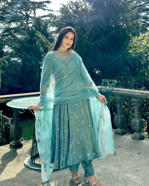 Woman in a light blue traditional outfit standing outdoors with greenery in the background
