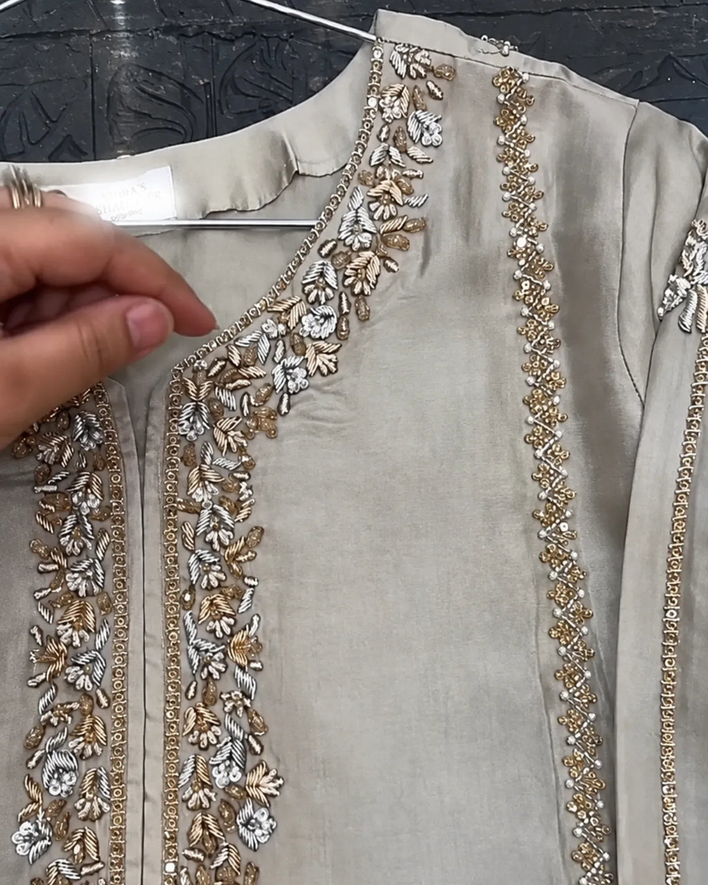 A close-up of a beige or light taupe kurta with ornate gold and silver floral embroidery along the neckline and front panels. A hand is visible holding the collar area, and the garment is shown on a hanger against a dark patterned background.