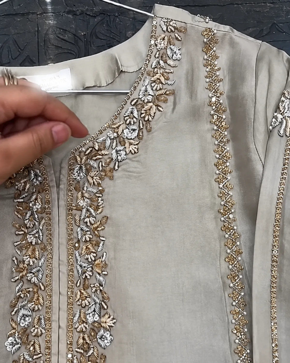 A close-up of a beige or light taupe kurta with ornate gold and silver floral embroidery along the neckline and front panels. A hand is visible holding the collar area, and the garment is shown on a hanger against a dark patterned background.