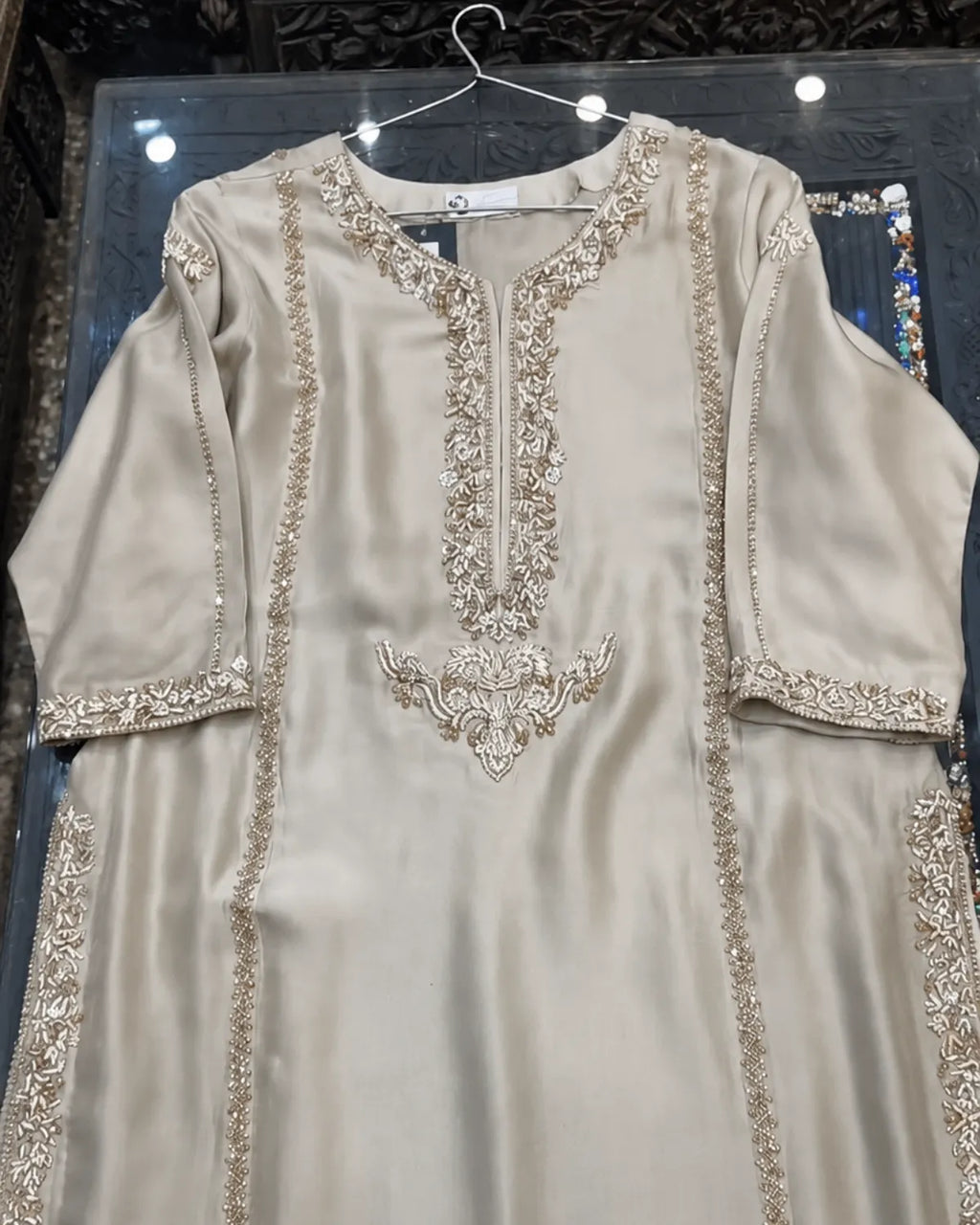 A beige satin kurta with intricate gold embroidery and mirror work, hanging on a wire hanger against a dark background.