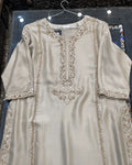 A beige satin kurta with intricate gold embroidery and mirror work, hanging on a wire hanger against a dark background.