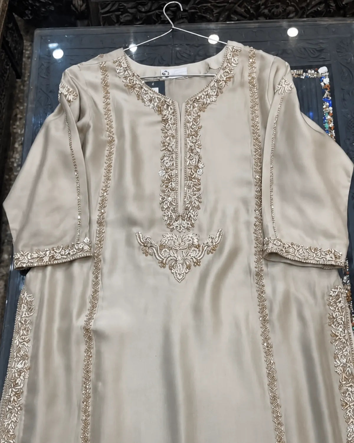 A beige satin kurta with intricate gold embroidery and mirror work, hanging on a wire hanger against a dark background.