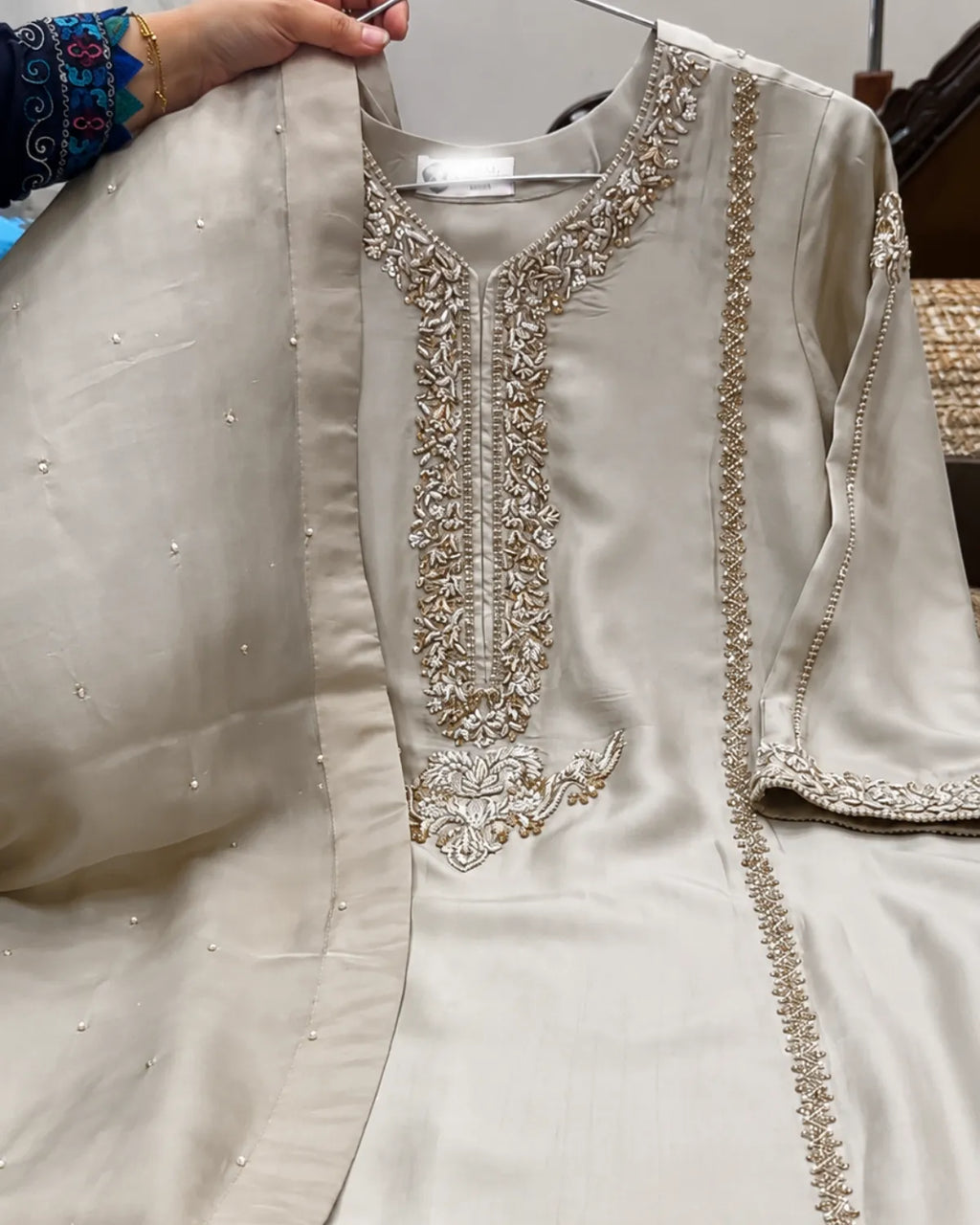 A light beige or cream-colored traditional embroidered kurta with a matching dupatta, held up by a hand on a hanger. The kurta features intricate metallic embroidery around the neckline, front placket, and sleeve cuffs.