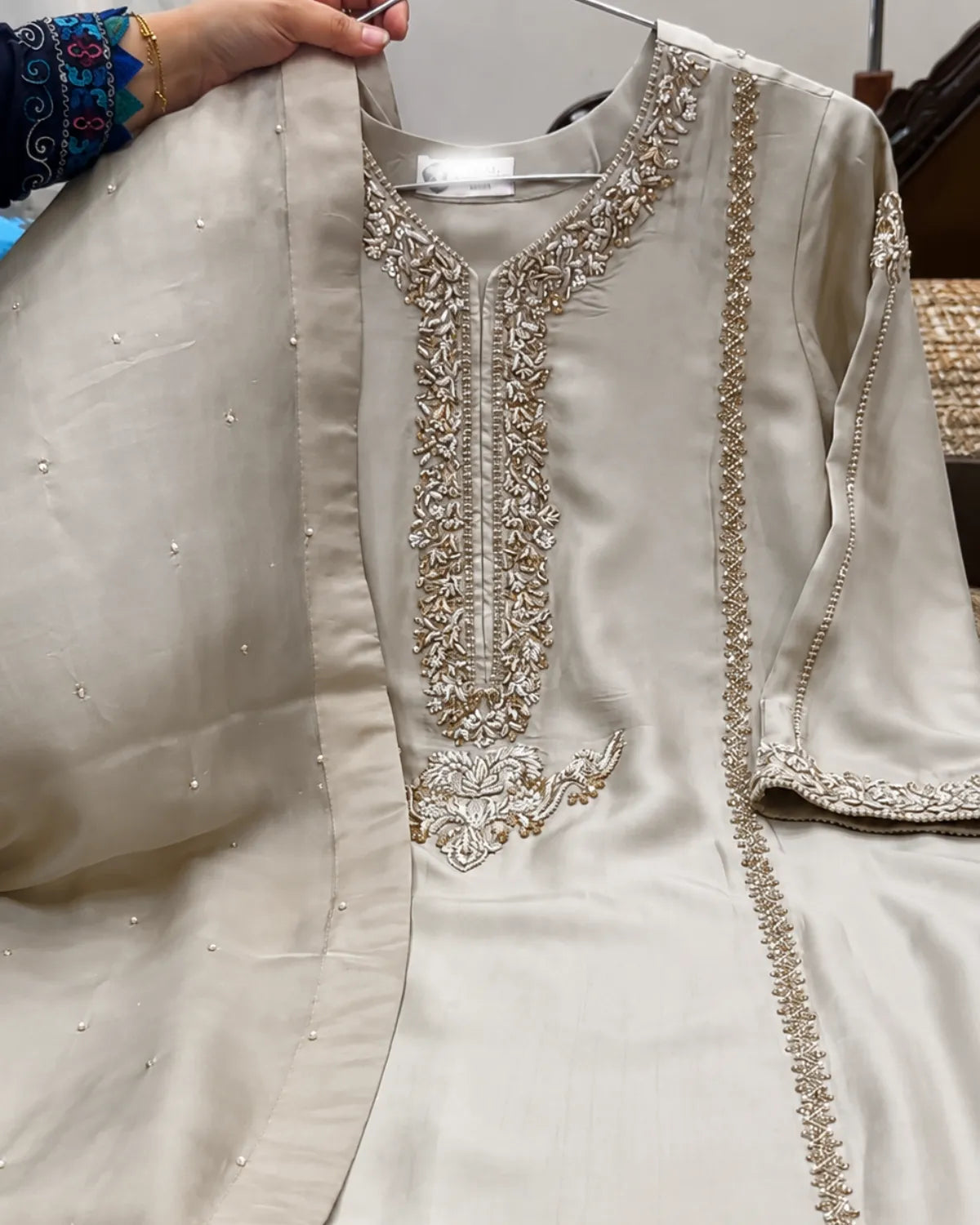 A light beige or cream-colored traditional embroidered kurta with a matching dupatta, held up by a hand on a hanger. The kurta features intricate metallic embroidery around the neckline, front placket, and sleeve cuffs.