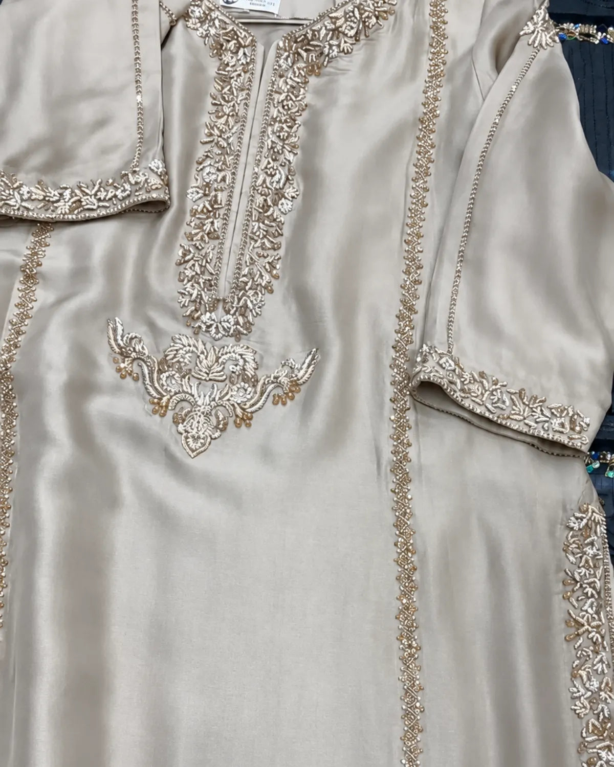 A close-up of a beige satin kurta featuring intricate gold and silver embroidery around the neckline, sleeve cuffs, and side borders.