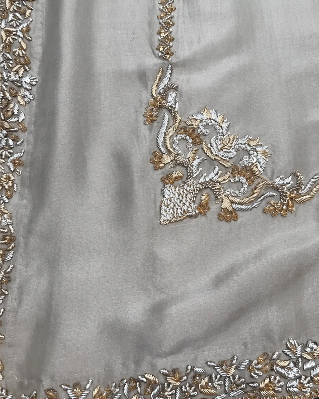 Close-up of a light gray fabric garment featuring intricate silver and gold floral embroidery along the border and neckline.