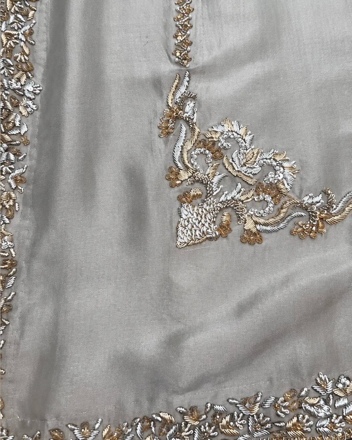 Close-up of a light gray fabric garment featuring intricate silver and gold floral embroidery along the border and neckline.