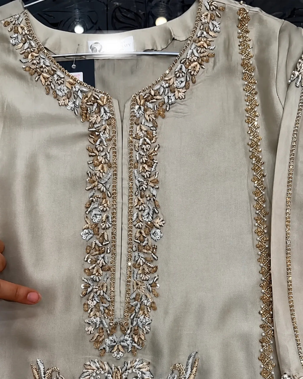A close-up of a beige or light khaki embroidered tunic top, likely a kurta, featuring ornate metallic gold and silver floral embroidery around the neckline and down the center front. A hand is visible on the left edge holding the fabric.