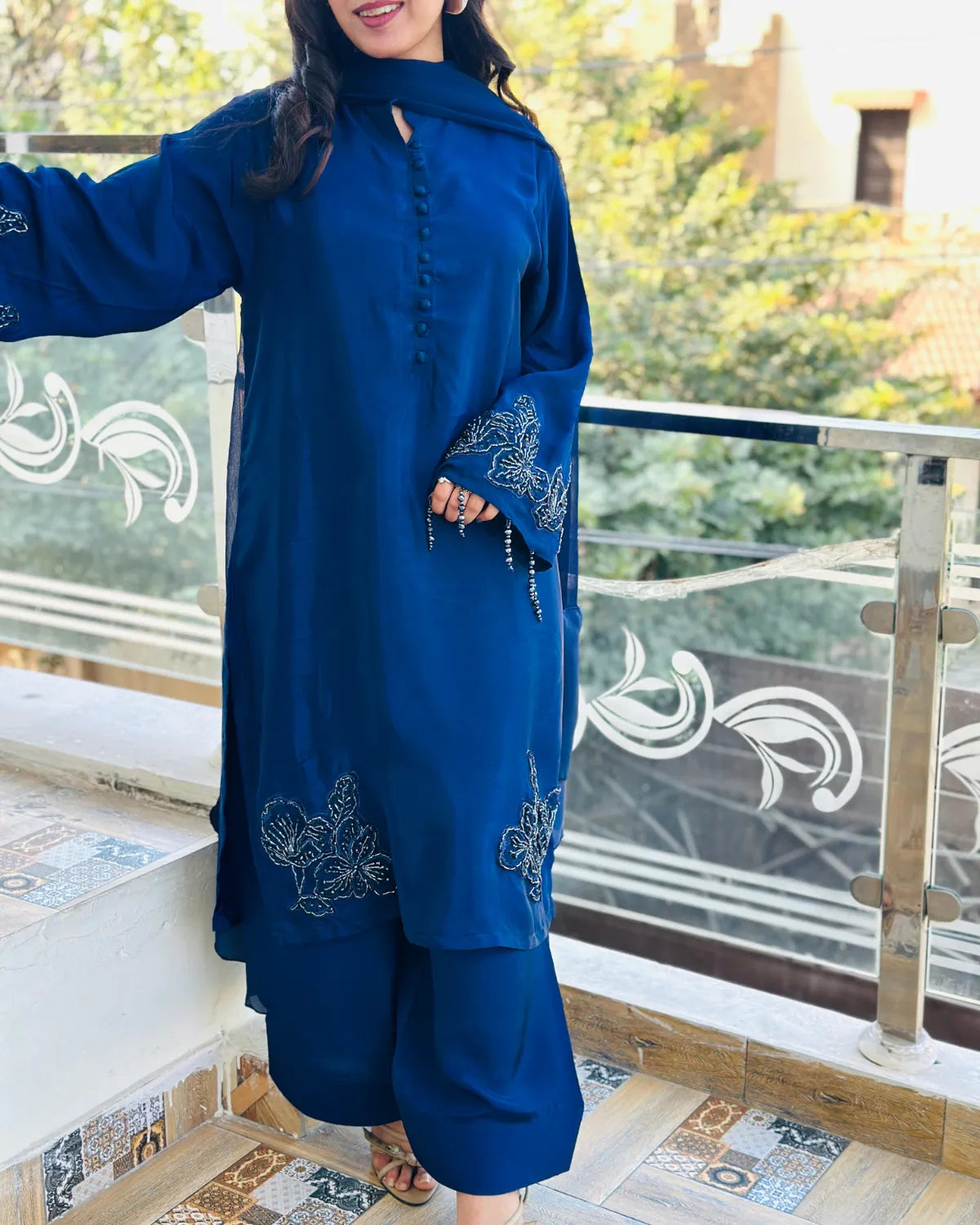 A woman wearing a royal blue traditional outfit with matching wide-leg pants, standing on an outdoor balcony with a glass railing and greenery in the background.
