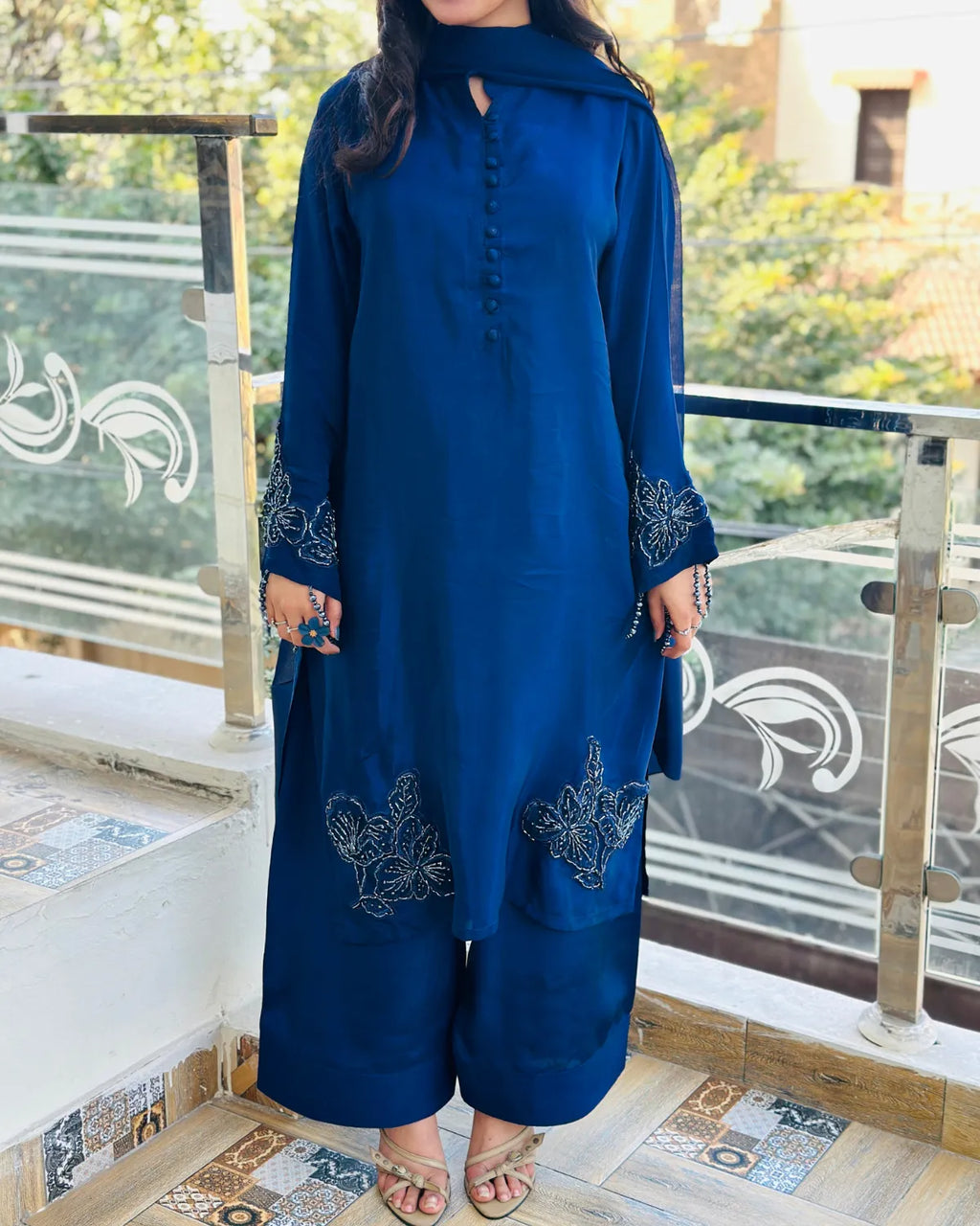 A woman wearing a deep blue traditional kurta set with matching wide-leg pants, featuring silver floral embroidery on the cuffs and lower front, standing on a tiled balcony.
