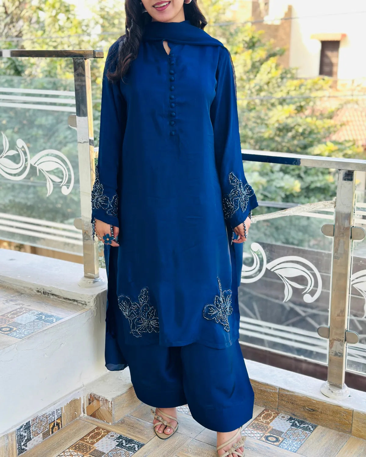 A woman wearing a royal blue kurta and trouser set with floral embroidery on a balcony. The outfit features a keyhole neckline, buttoned front, and wide-leg pants.