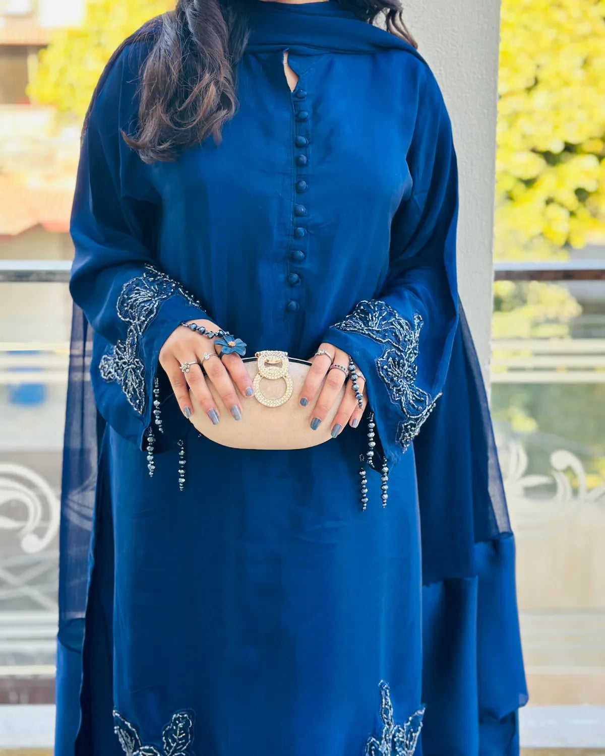 A woman wearing a royal blue embroidered abaya dress with a matching sheer dupatta and holding a beige clutch bag.