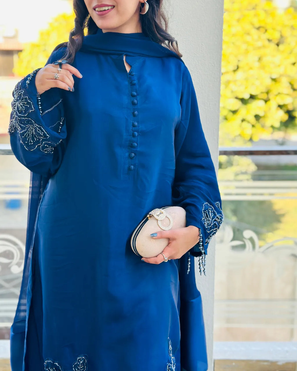 A woman wearing a royal blue long kurta with silver embroidery on the sleeves and a matching scarf, holding a cream clutch bag.