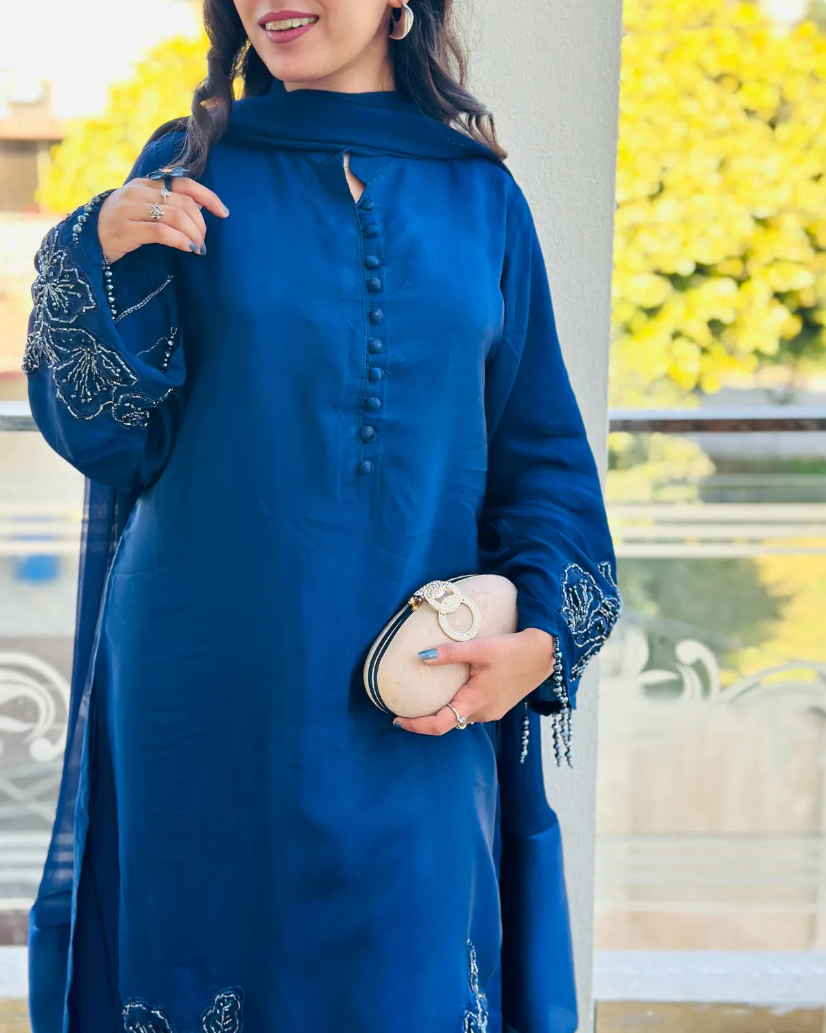 A woman wearing a royal blue long kurta with silver embroidery on the sleeves and a matching scarf, holding a cream clutch bag.
