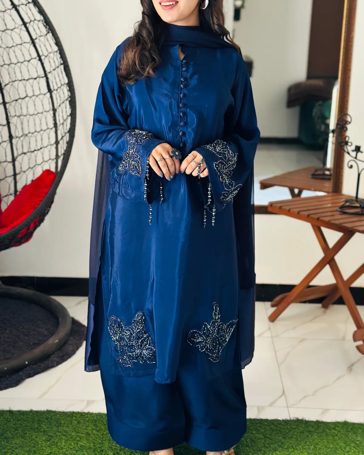 A woman wearing a deep blue traditional outfit with matching pants and a draped scarf, standing indoors. The outfit has silver embroidered details on the sleeves and lower front, and she is holding black prayer beads.
