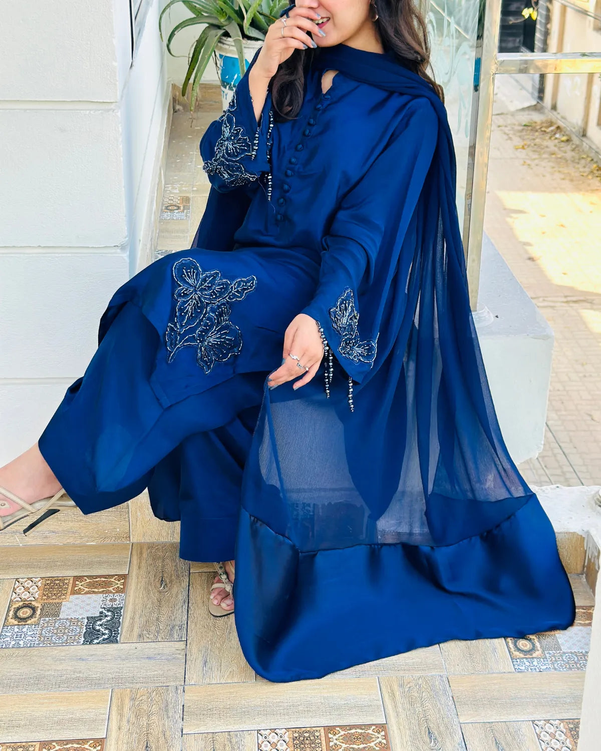 A woman wearing a royal blue traditional outfit with wide-leg pants, embroidered sleeves, and a sheer draped overlay, seated on a tiled outdoor step.