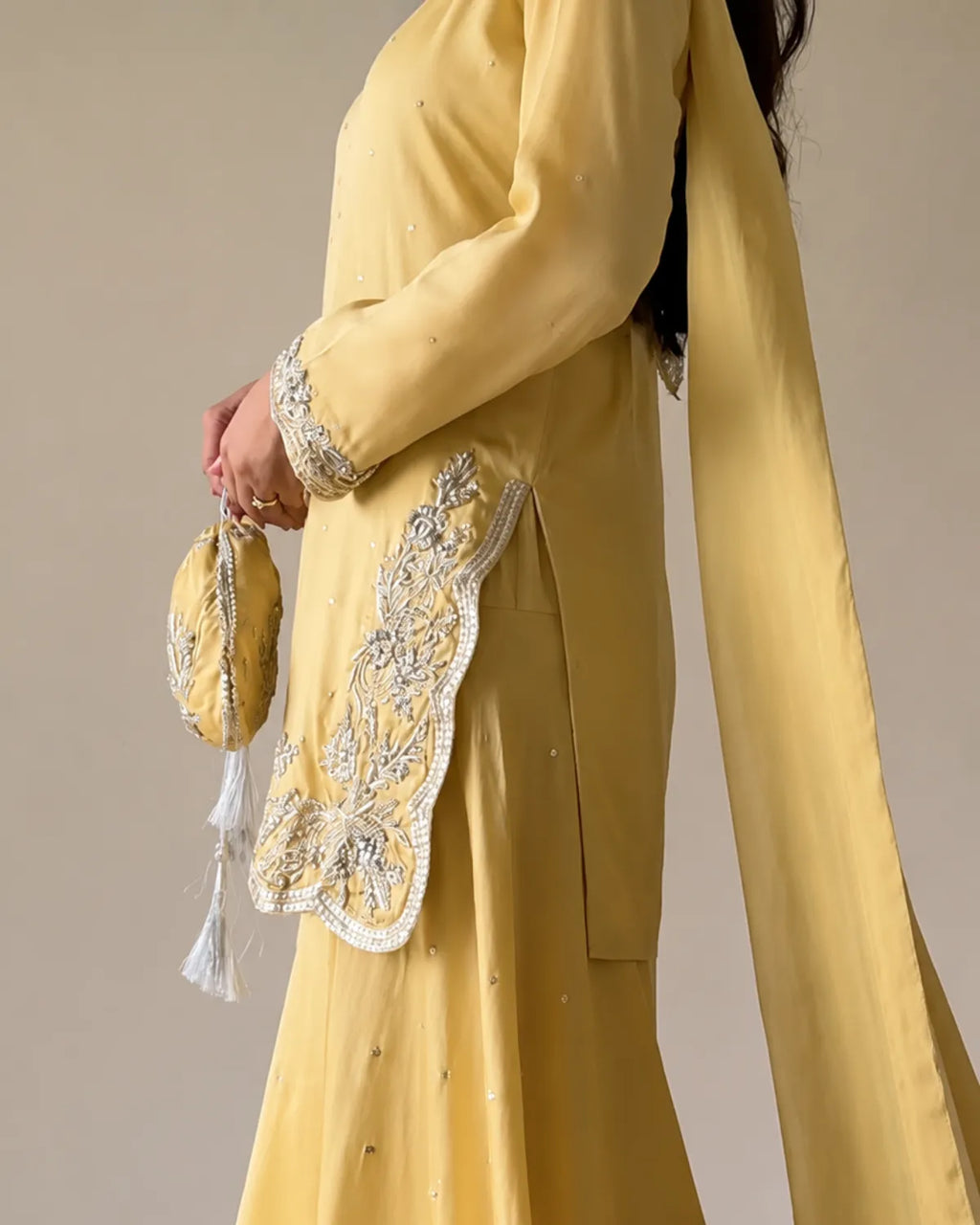 A person wearing a soft yellow traditional outfit with silver embroidered detailing on the sleeve and hem, holding a matching yellow embroidered clutch bag with white tassels against a plain beige studio background.