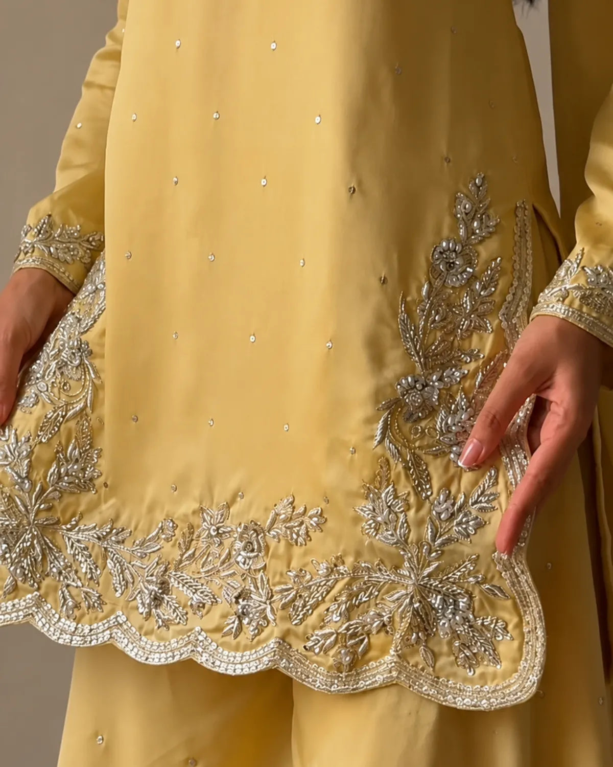 A close-up of a yellow traditional outfit with intricate silver embroidery and mirror work along the hem and cuffs, worn by a model whose hands are visible.