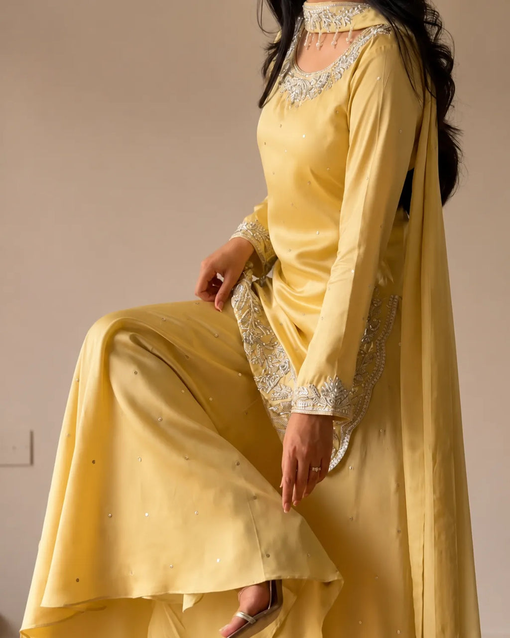 A woman wearing a yellow traditional outfit with silver embroidered details and a matching sheer dupatta, posed against a plain beige wall.