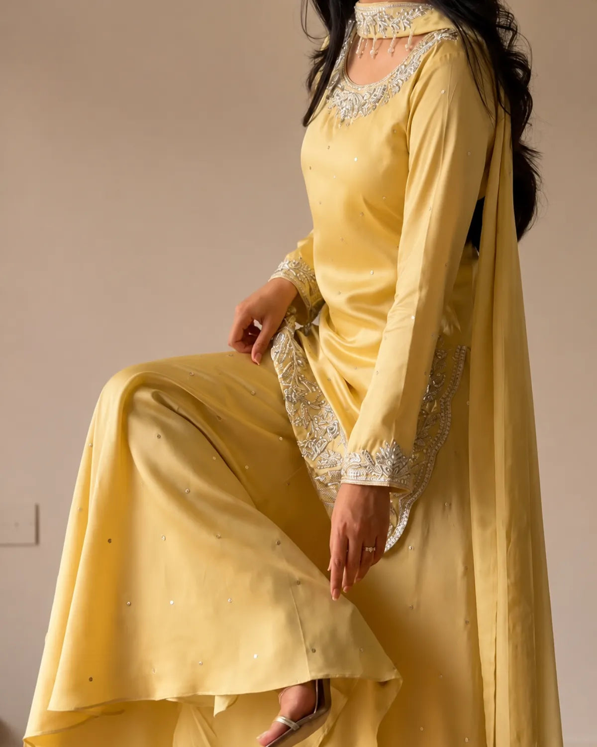 A woman wearing a yellow traditional outfit with silver embroidered details and a matching sheer dupatta, posed against a plain beige wall.
