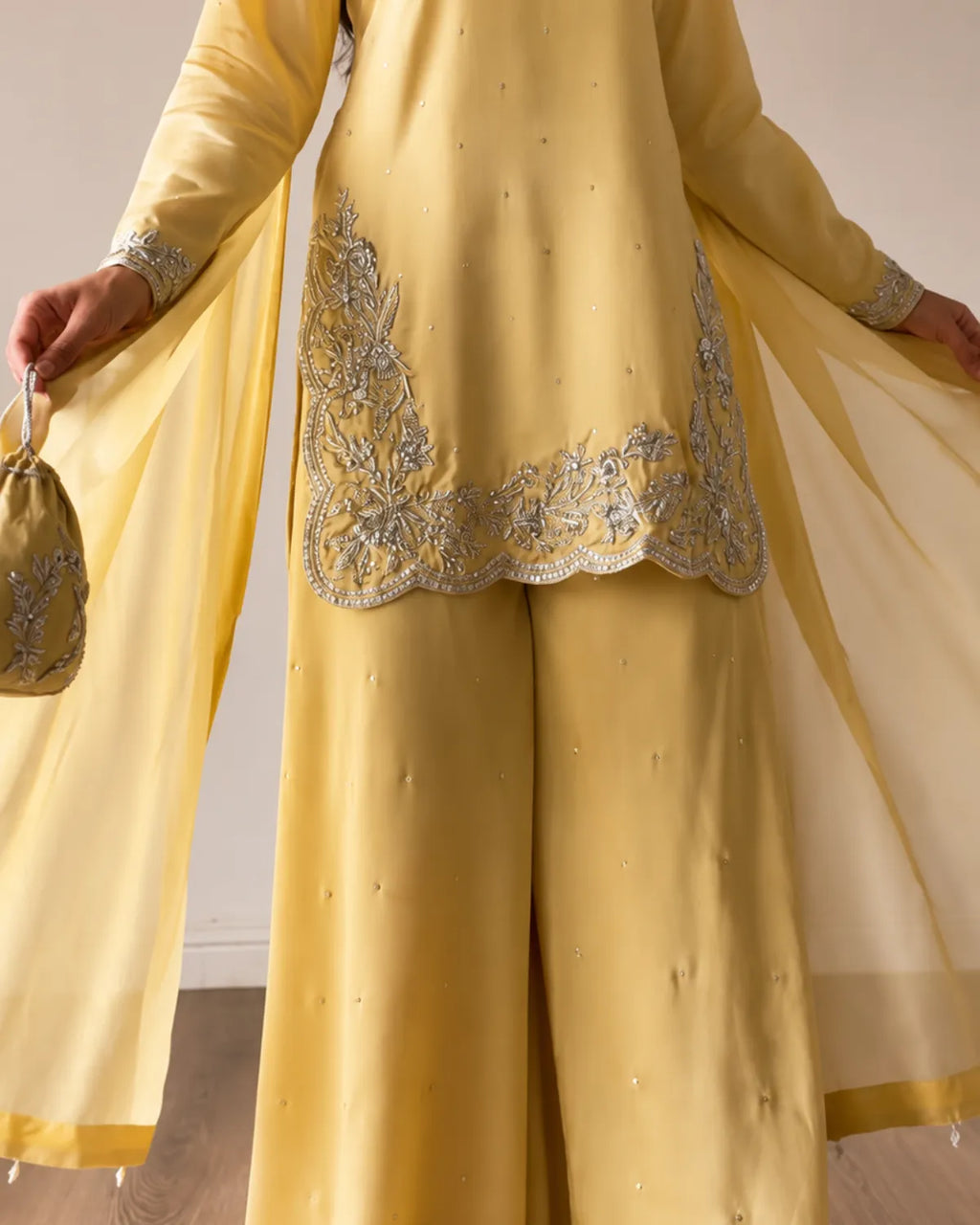 A person wearing a yellow traditional outfit with wide-leg pants, a matching sheer drape, and silver embroidered detailing on the front, holding a small matching yellow pouch.