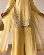 A person wearing a yellow traditional outfit with wide-leg pants, a matching sheer drape, and silver embroidered detailing on the front, holding a small matching yellow pouch.